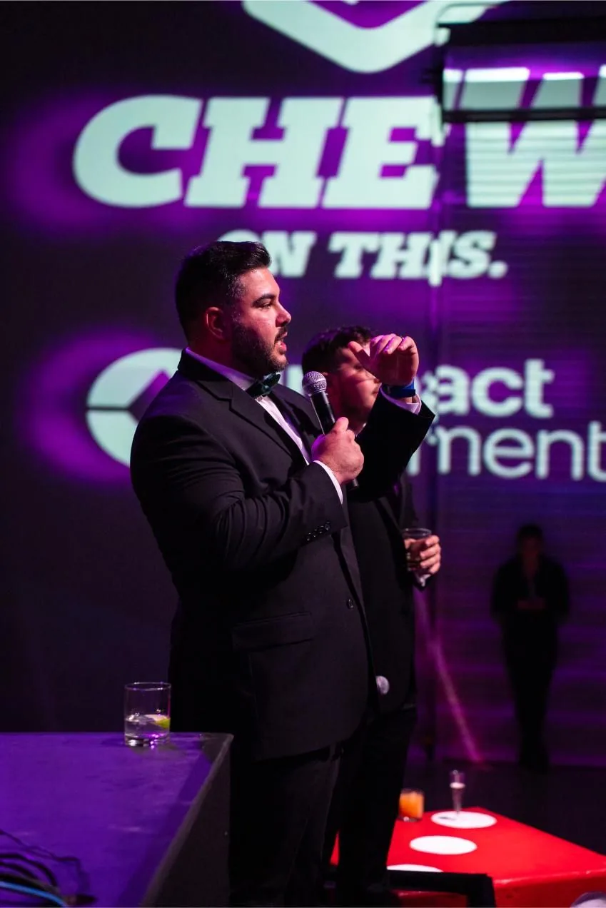 Host speaking on stage with a microphone at Bad After Dark in Las Vegas, partner logos projected behind him