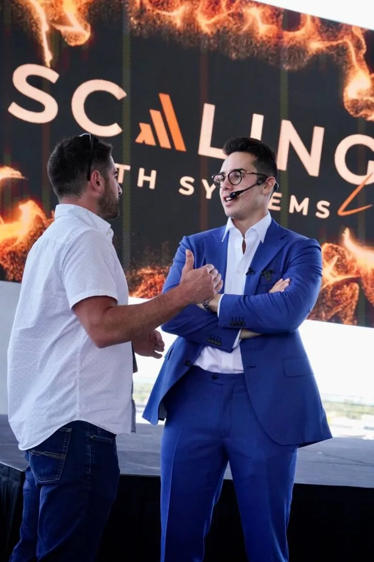 Two men laughing in front of the Scaling With Systems screen at Scaling With Systems LIVE in Miami